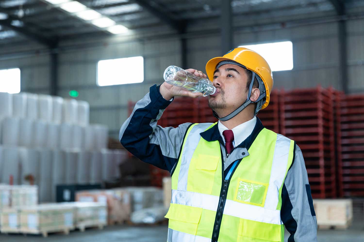 Warehouse worker in an orange hard hat and yellow hi-vis vest drinking from a water bottle, with red pallet racks behind him