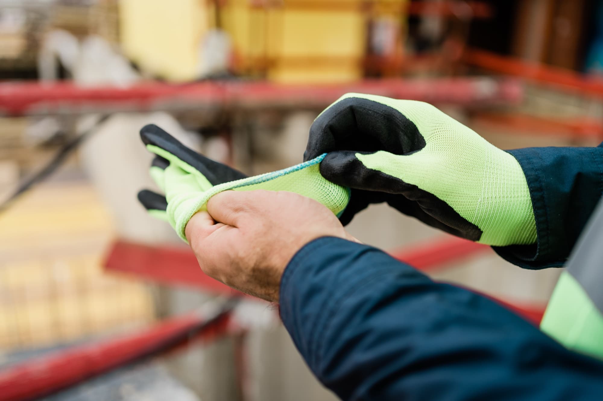 Worker in a dark jacket pulling on high-visibility green work gloves at a construction site with scaffolding in the background
