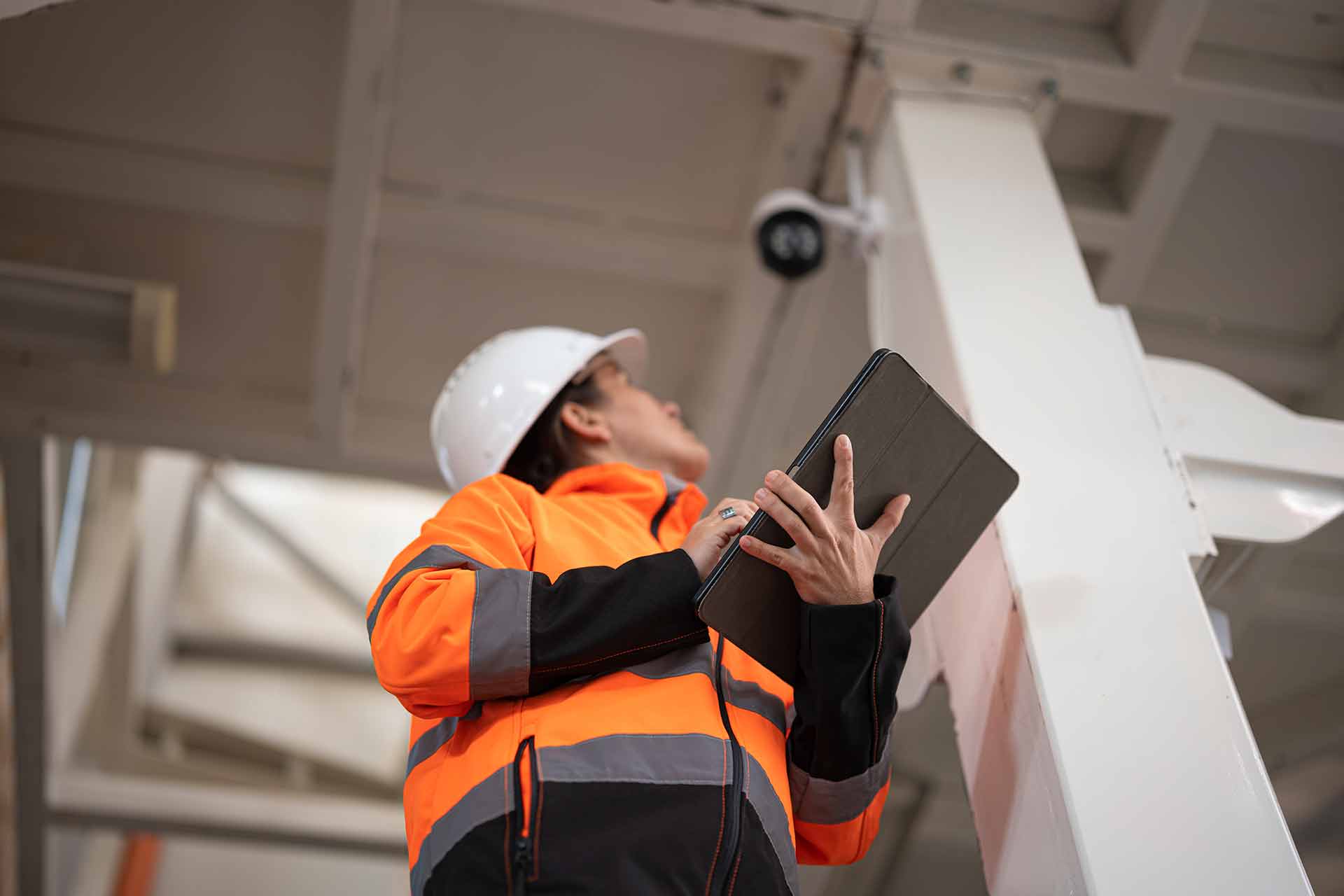Worker in a white hard hat and orange hi-vis jacket holding a tablet while looking up at a device mounted on a white steel beam