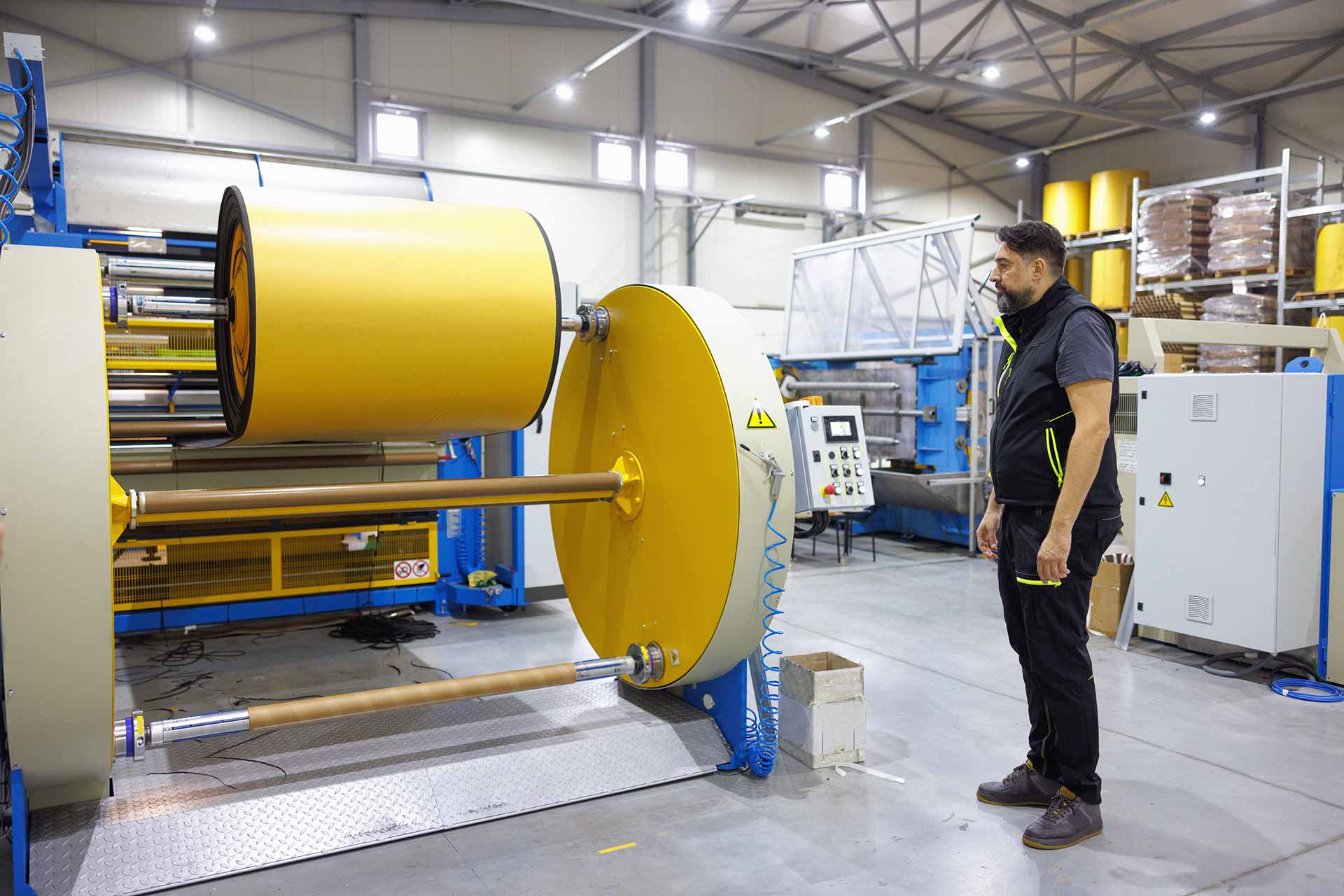 Worker in a black vest watching a large yellow roll of material on an unwinding machine in a brightly lit production facility