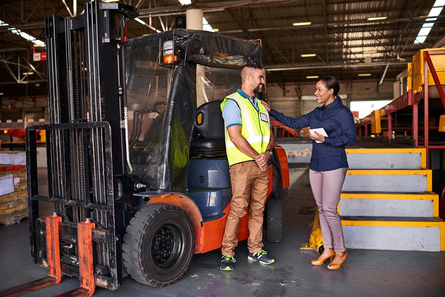 Woman holding a tablet resting a hand on the shoulder of a smiling forklift operator in a hi-vis vest beside his forklift