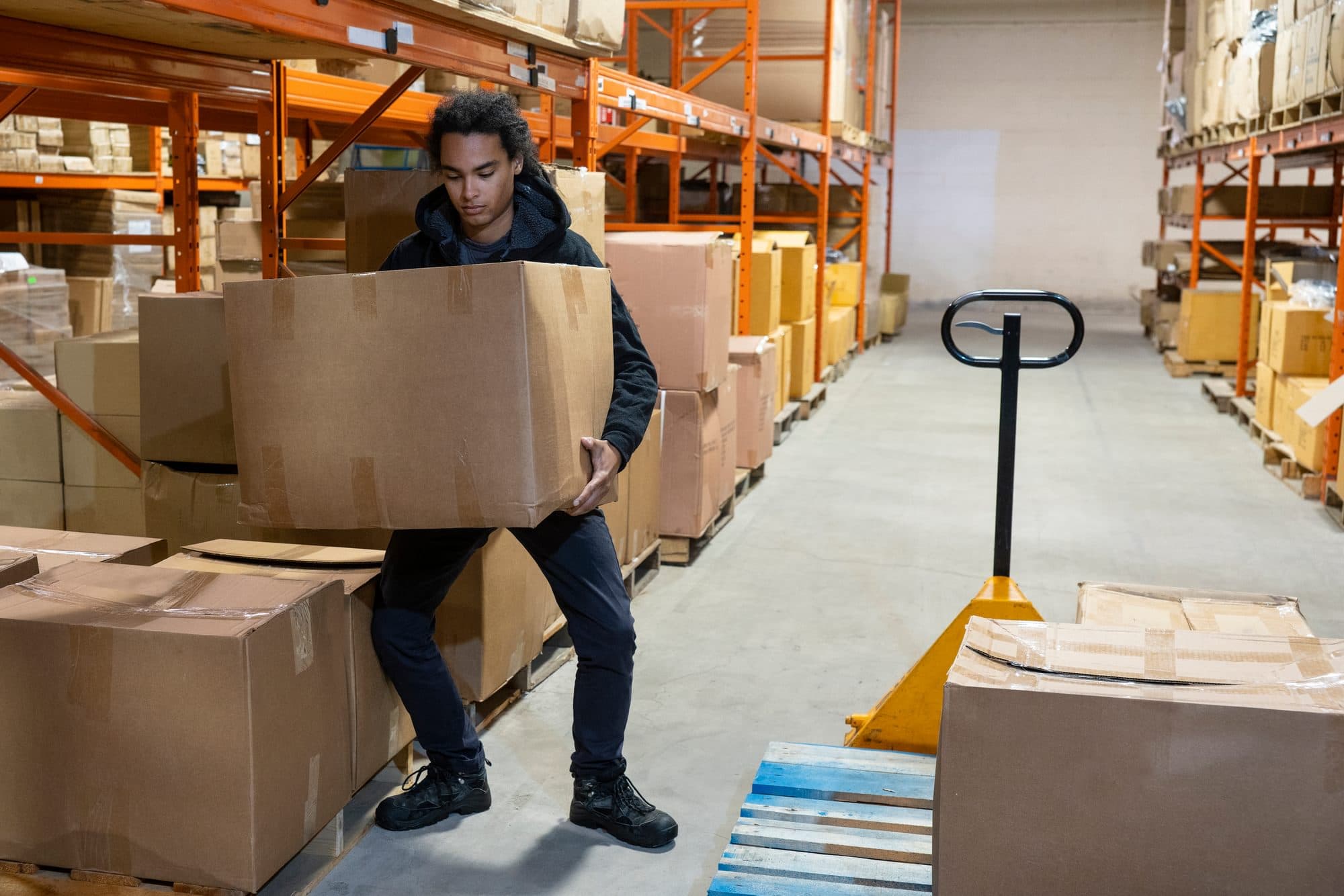 Warehouse worker carrying a large cardboard box between storage racks.