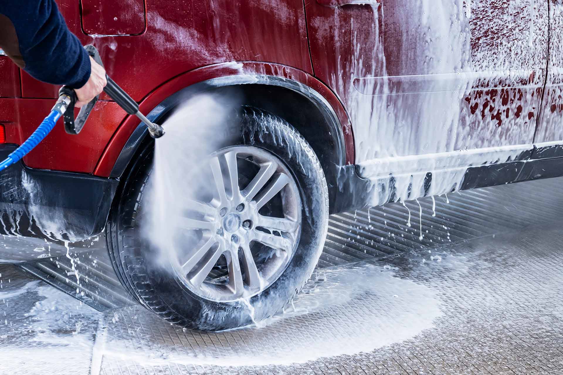 Worker spraying foam from a pressure washer onto the wheel of a red SUV at a car wash