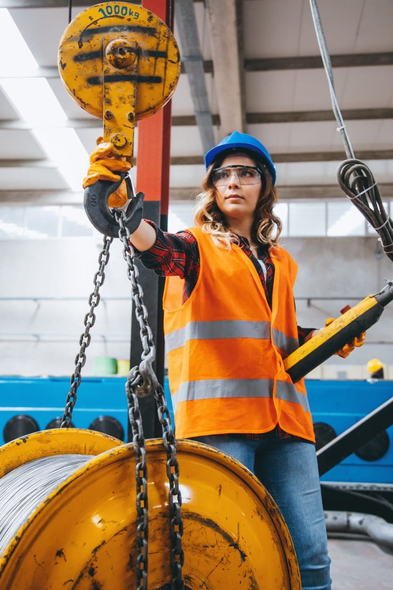 Worker in a hard hat, safety glasses and high-visibility vest guiding an overhead crane hook onto a load